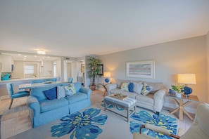 A modern living room with a blue sofa, a white armchair, and a coffee table. There is a dining area with a blue table and chairs, and a kitchen in the background. - 201 Tidewater in Ponce Inlet, FL (Ponce Inlet)