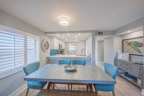 A modern kitchen with a central island, a dining area with blue chairs, and a large window with blinds. - 201 Tidewater in Ponce Inlet, FL (Ponce Inlet)