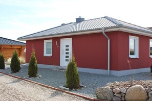 A red house with a grey roof, white door, and windows, surrounded by gravel and small trees. - Holiday home Sandseele (Fehmarn)