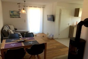 A living room with a sofa, a dining table with chairs, a TV mounted on the wall, and a fireplace. - Holiday home Sandseele (Fehmarn)