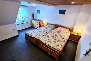 Ein Schlafzimmer mit einem hölzernen Bett, einem Schminktisch, einem Nachttisch, einem Fenster mit Vorhängen und einer Heizung.