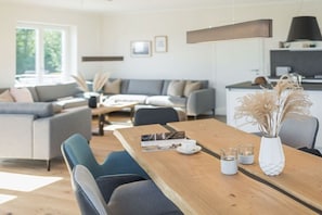 A modern living room with a wooden dining table, grey sofas, and a kitchen area in the background. - Single-family house Sundseele No. 14 (Fehmarn)