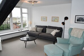 A living room with a sofa, a chair, a coffee table, and a radiator. There are two windows and a picture frame on the wall. - Holiday apartment with 2 bedrooms (Fehmarn)