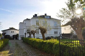 A white building with a cobblestone path leading to the entrance, surrounded by greenery and a metal fence. - Holiday apartment with 2 bedrooms (Fehmarn)