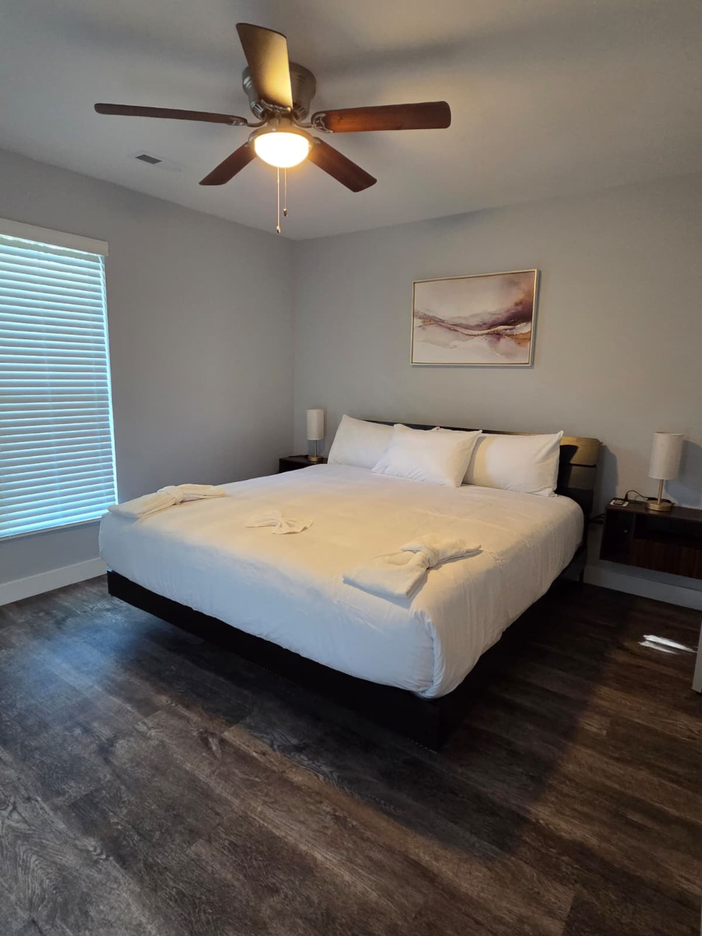 A bedroom with a bed, a ceiling fan, a nightstand, a lamp, and a painting on the wall.