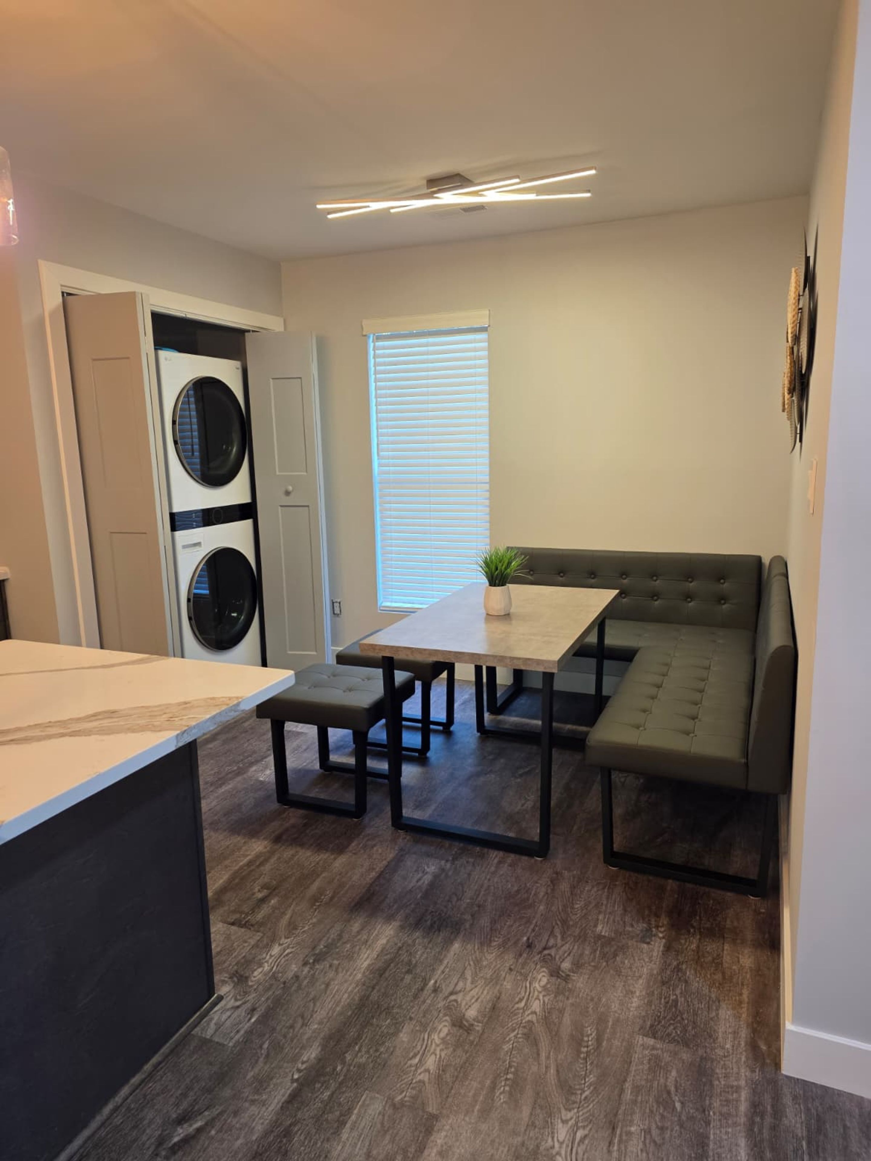 A compact living space with a kitchenette, a dining area with a table and bench, a built-in washer and dryer, and a large window with blinds.