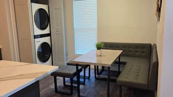 A compact living space with a kitchenette, a dining area with a table and bench, a built-in washer and dryer, and a large window with blinds.