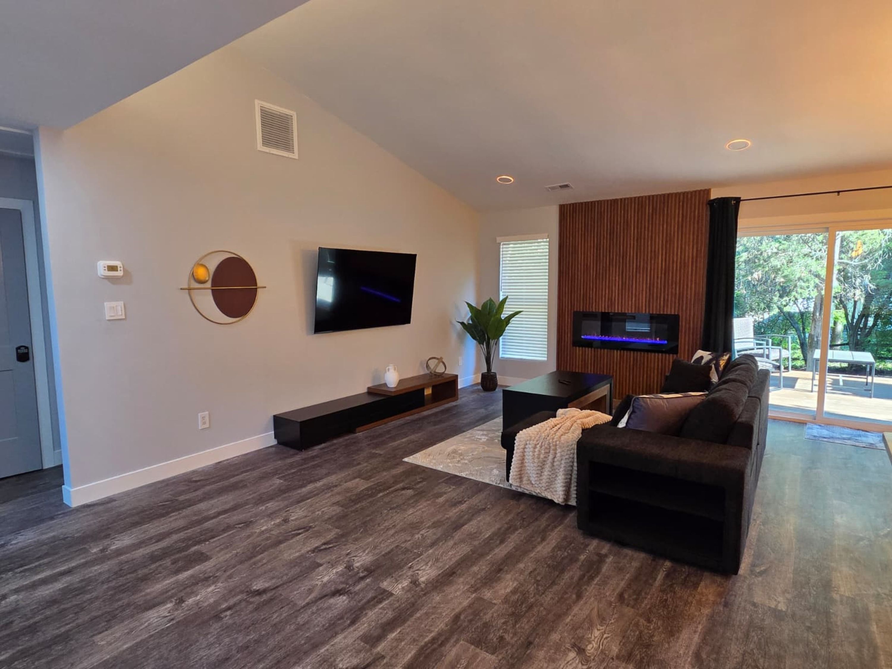 A modern living room with a flat-screen TV, dark wood flooring, a sectional sofa, and a large window offering a view of the outdoors.