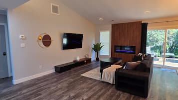 A modern living room with a flat-screen TV, dark wood flooring, a sectional sofa, and a large window offering a view of the outdoors.
