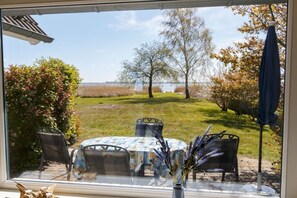 A patio with a table and chairs, a vase with flowers, and a view of a grassy area with trees and a body of water. - Holiday home with 2 bedrooms (Born a. Darß)