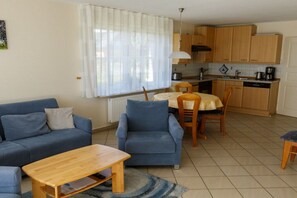A compact living space with a sofa, a dining table, and chairs, a kitchen area with cabinets and appliances, and a window with curtains. - Holiday home with 2 bedrooms (Born a. Darß)