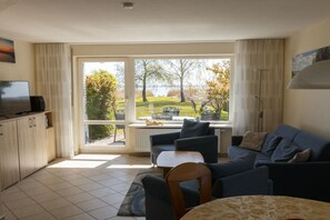 A living room with a television, seating, and a view of a garden and lake. - Holiday home with 2 bedrooms (Born a. Darß)