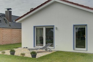 A single-story house with a tiled patio, a lawn, and a lounge chair. - Holiday home with 1 bedroom (Fehmarn OT Wulfen)