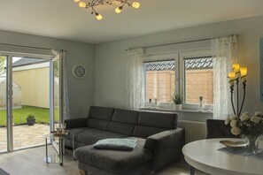 A modern living room with a sofa, a dining table, and a window with curtains. - Holiday home with 1 bedroom (Fehmarn OT Wulfen)