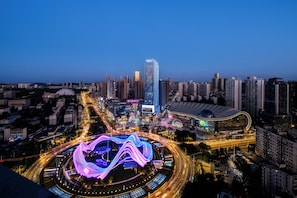 Paisagem urbana ao entardecer, com edifícios iluminados, uma estrutura curva imponente e um ambiente urbano denso.