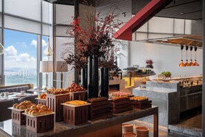 A buffet setup with various food items, a large decorative plant, and a city view through large windows.