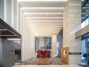 A modern hotel lobby with a central reception desk, a patterned floor, and a high ceiling with linear lighting.