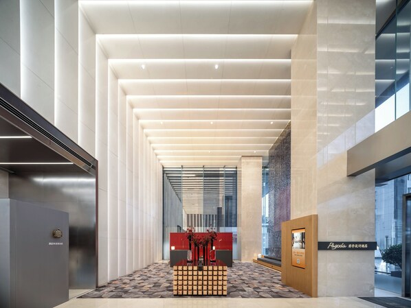 A modern hotel lobby with a central reception desk, a patterned floor, and a high ceiling with linear lighting.