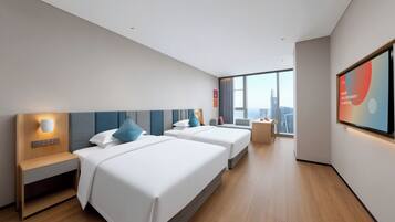A hotel room with two beds, a desk, a chair, a large window with a city view, and a flat-screen TV mounted on the wall.