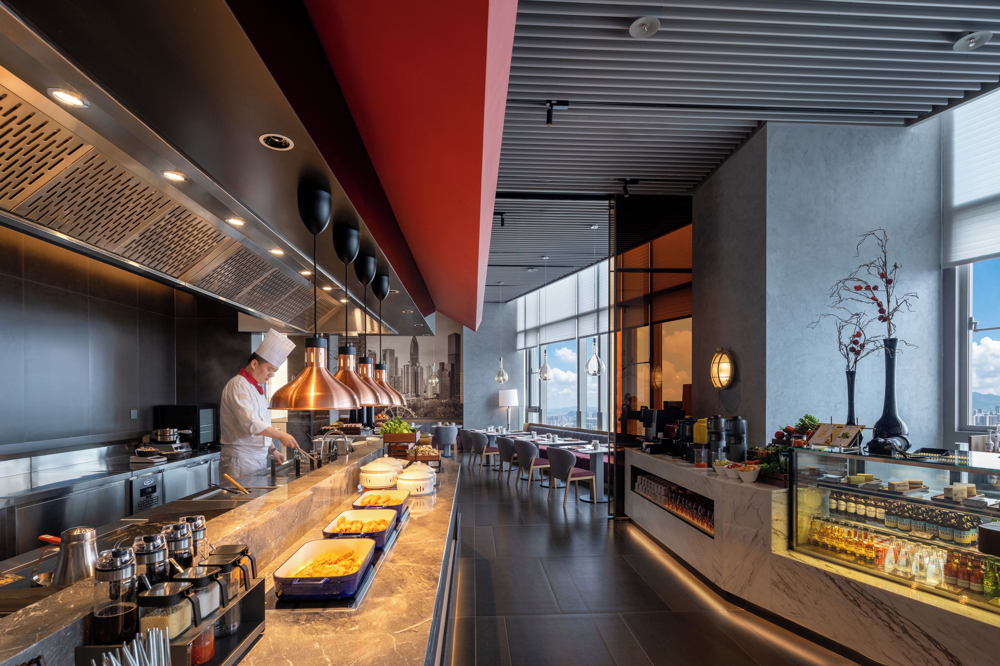 A modern restaurant with a chef preparing food, a buffet with various dishes, and a view of the city through large windows.
