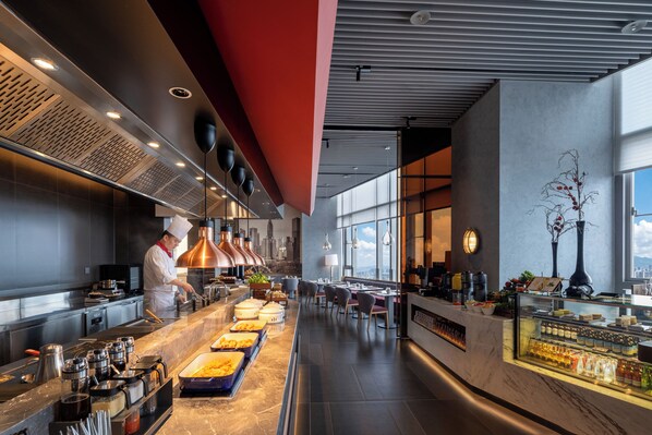 A modern restaurant with a chef preparing food, a buffet with various dishes, and a view of the city through large windows.