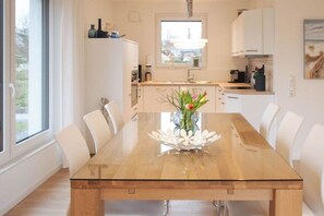 A modern dining area with a glass-top table, white chairs, and a vase with flowers. - Holiday home with 3 bedrooms (Timmendorfer Strand)