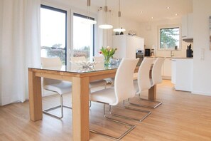 A modern dining area with a glass table, white chairs, and pendant lighting. - Holiday home with 3 bedrooms (Timmendorfer Strand)