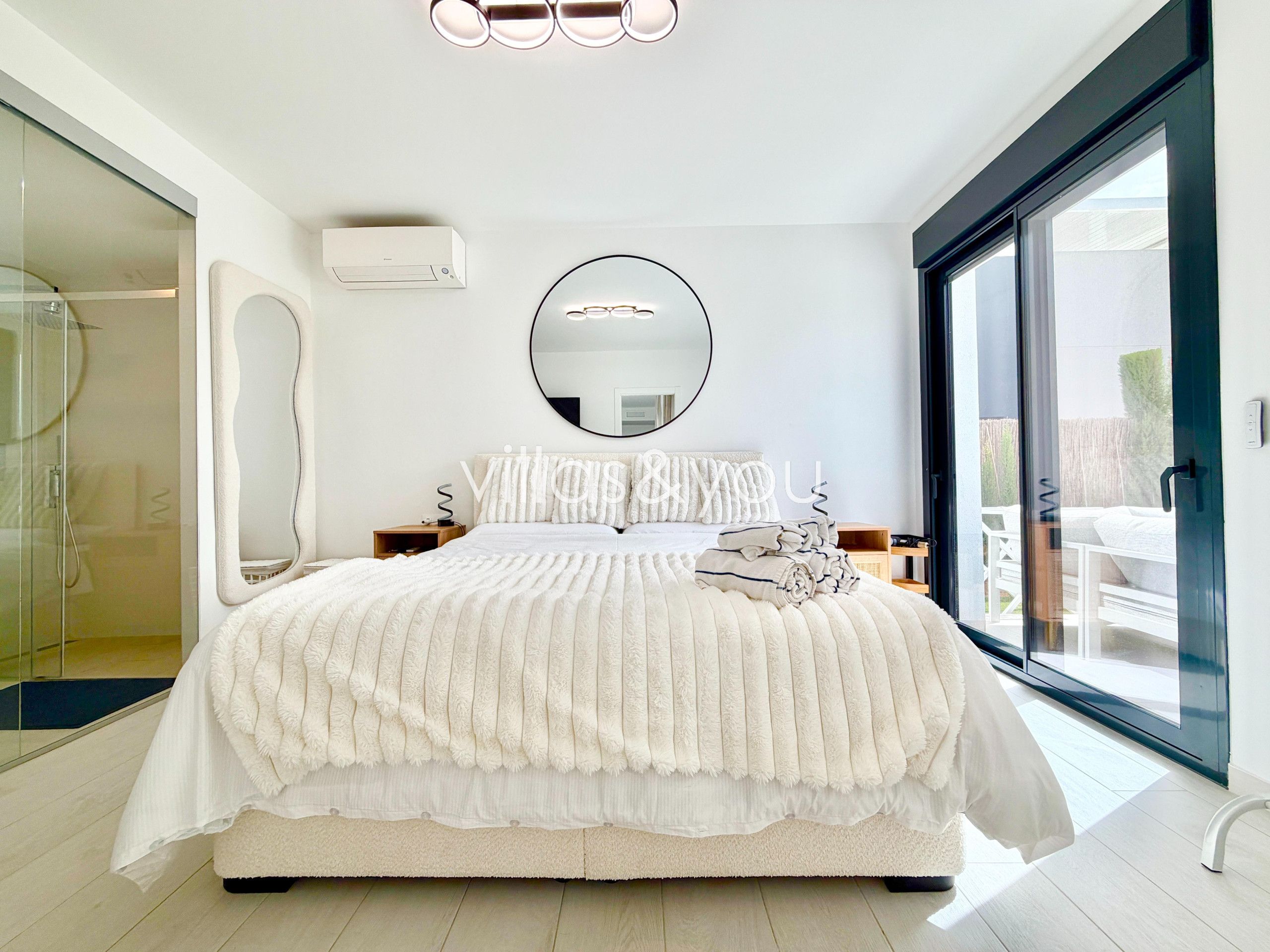A modern bedroom with a large bed, a round mirror, and a sliding glass door leading to an outdoor area.