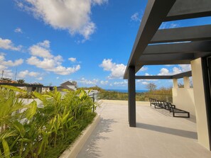 Ein überdachter Außenbereich mit Blick auf das Meer, eine Sitzecke und ein Garten mit tropischen Pflanzen.