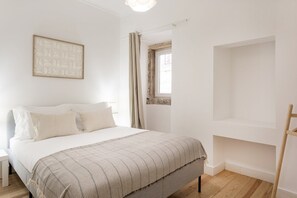 Ein Schlafzimmer mit einem Bett, einem Nachttisch, einem Fenster mit Vorhängen und einem an der Wand befestigten Kunstwerk.