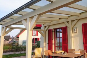 Une terrasse extérieure avec une table et des chaises, un espace couvert avec un toit, et des volets rouges sur le bâtiment.