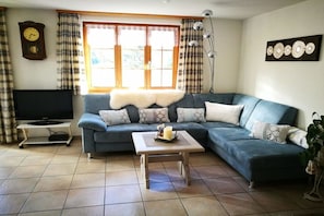Ein Wohnzimmer mit einem Ecksofa, einem Flachbildfernseher auf einem Schrank, einem kleinen Tisch mit einer Kerze und einem großen Fenster mit Vorhängen.