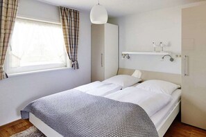 A bedroom with a bed, a wardrobe, a window with curtains, a hanging light fixture, and a shelf with decorative items. - Holiday home with 3 bedrooms (Timmendorfer Strand)