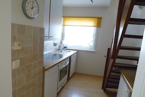 A compact kitchen with a sink, stove, and oven, a window with a yellow blind, and a clock on the wall. - Holiday home with 3 bedrooms (Timmendorfer Strand)