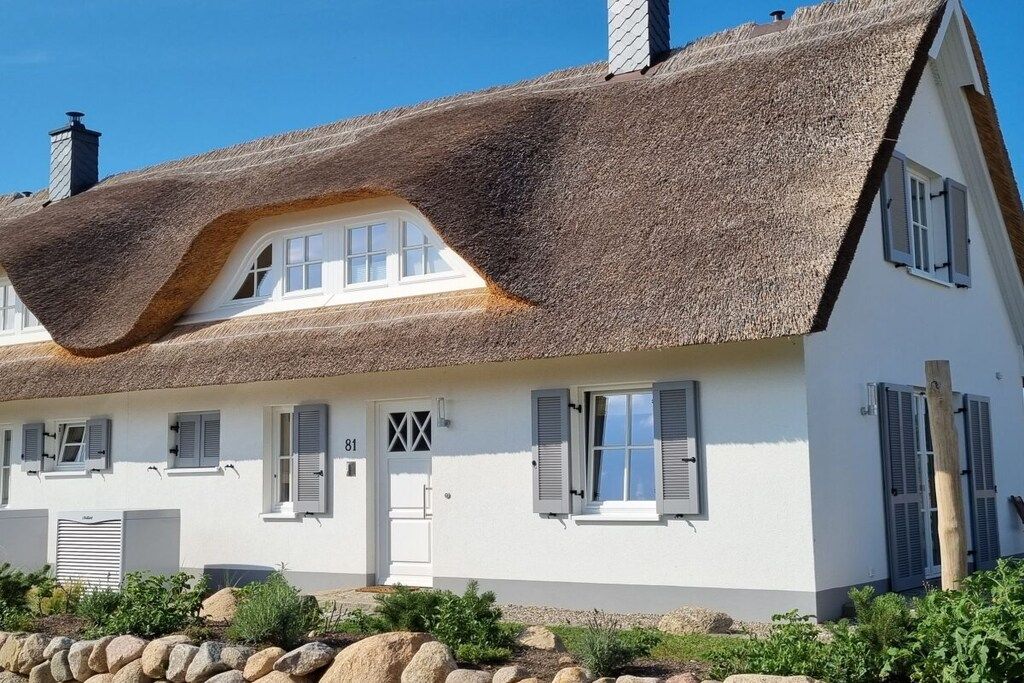 A white house with a thatched roof, a chimney, and a stone wall in front.
