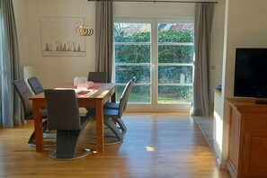 A dining area with a table and chairs, a television on the right, and a large window with curtains. - Holiday home with 4 bedrooms (Ahrenshoop)