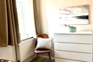 Una habitación con una silla, un radiador, un aparador blanco, una ventana con cortinas y un cuadro colgado en la pared.