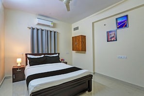 Ein Hotelzimmer mit einem Bett, einem Nachttisch, einer Klimaanlage, einem Deckenventilator, einem Wäscheschrank und zwei gerahmten Bildern an der Wand.