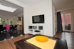 Ein modernes Wohnzimmer mit Flachbildfernseher an der Wand, einem Esstisch mit Stühlen und einer Küche im Hintergrund.