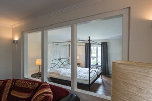 Ein Schlafzimmer mit Himmelbett, Nachttisch, Lampe und einem großen Fenster mit Vorhängen.