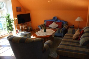 Ein gemütliches Wohnzimmer mit einer Couch, Sesseln, einem Couchtisch mit weißer Tischdecke, einem Fernseher und einer Pflanze.