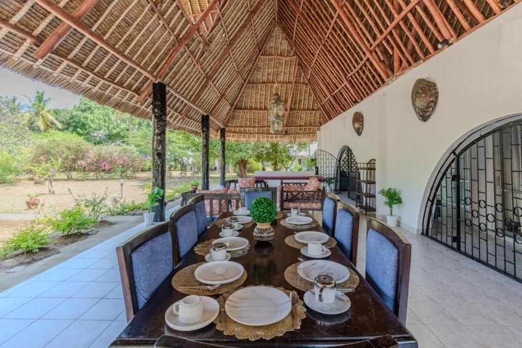 Un comedor con techo de paja, una mesa larga dispuesta para una comida, y vistas del jardín circundante.