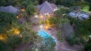 Eine Vogelperspektive zeigt ein Resort mit einem Swimmingpool, umgeben von üppiger Vegetation und mehreren strohgedeckten Gebäuden.