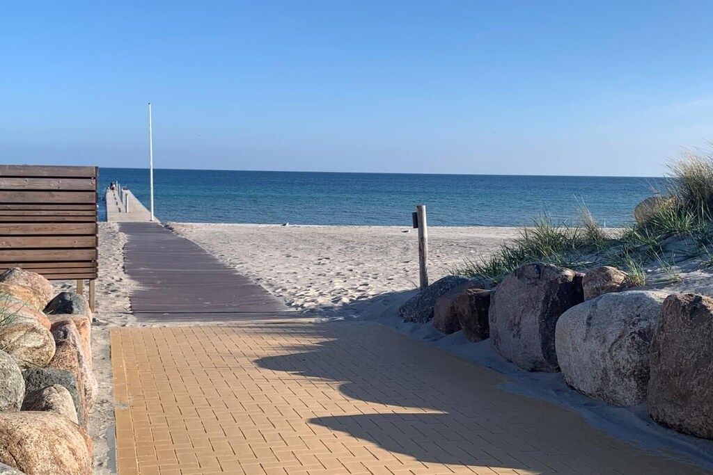 Een verhard pad dat naar een strand leidt, met een houten constructie aan de linkerkant en grote rotsen aan de rechterkant.