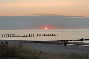 Ein Strand mit Sonnenuntergang, Menschen die spazieren gehen und einem hölzernen Steg.