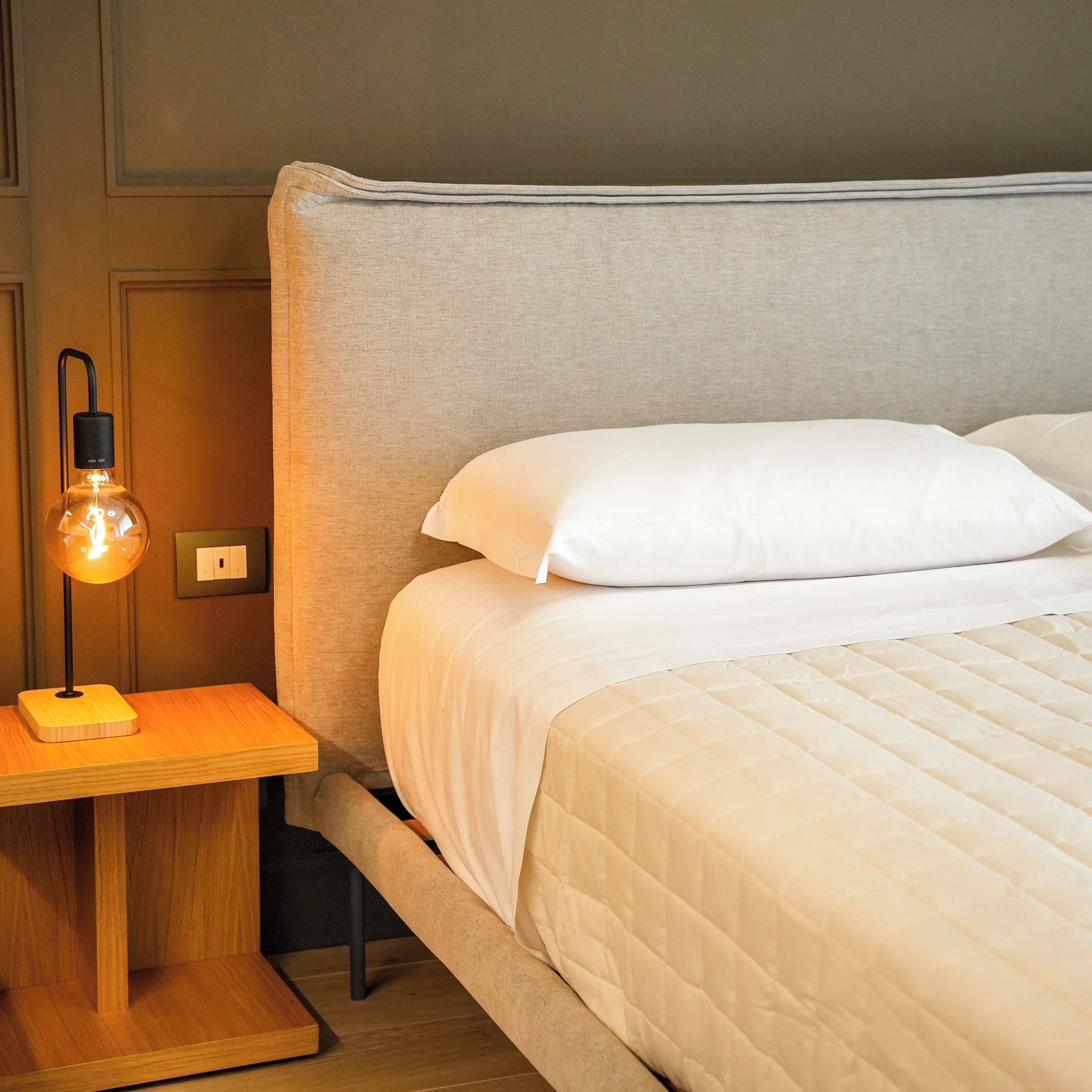 Ein ordentlich bezogenes Bett mit weißer Bettwäsche, eine Nachttischlampe mit schwarzem Fuß und ein Holznachttisch.