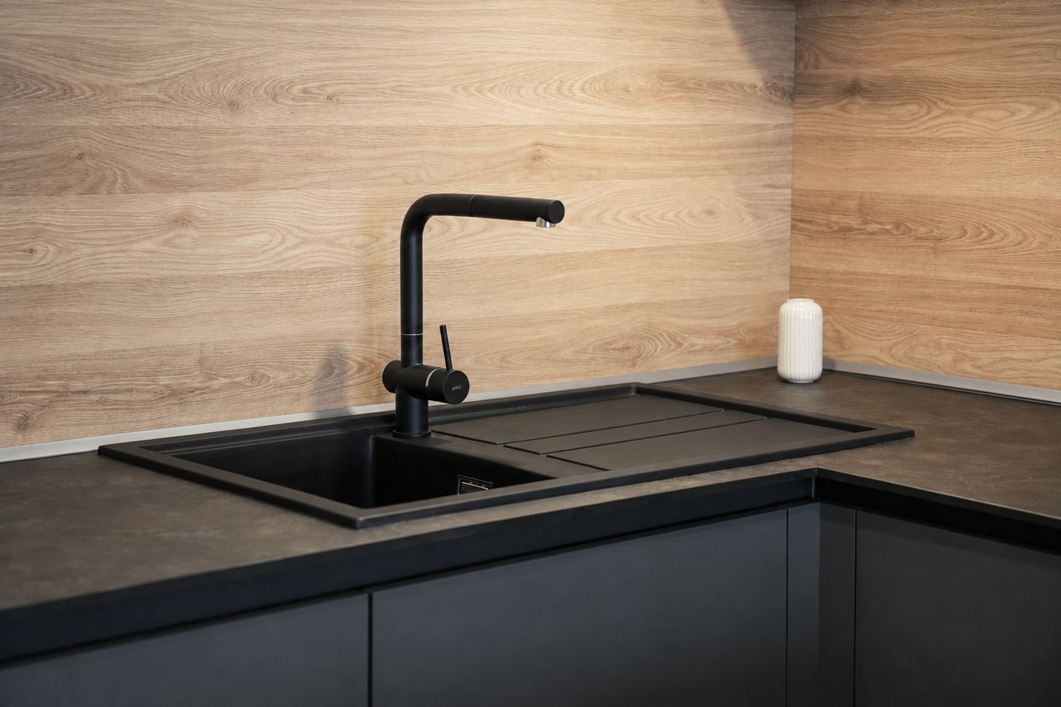 Una cocina con un lavamanos negro, un protector contra salpicaduras de madera y un recipiente blanco sobre la encimera.