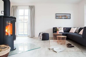A modern living room with a wood-burning stove, a dark sofa, a glass coffee table, and a view of the outdoors through a window. - ||------------------------Reed Warbler 45---------------------|| (Fuhlendorf)