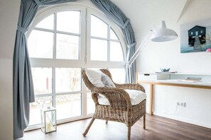 A room with a large arched window, a wicker chair with a cushion, a white desk with a lamp, and a painting on the wall. - ||------------------------Reed Warbler 45---------------------|| (Fuhlendorf)