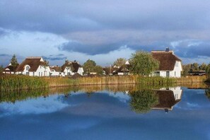 Reihenweise weiße Häuser mit Strohdächern an einem ruhigen See, der die Gebäude und den Himmel spiegelt.